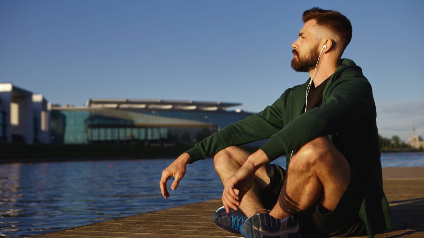 Bearded man in a green hoodie sits on a wooden dock by the water, wearing earbuds and looking to the right against a clear blue sky.