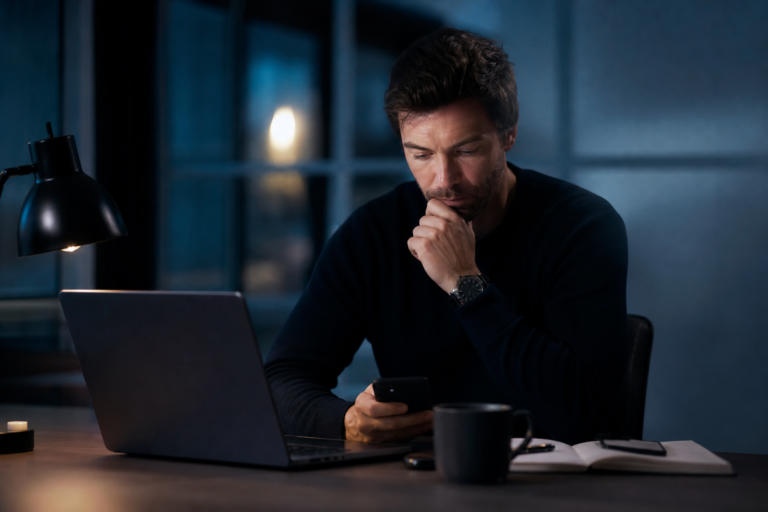 Man in a dark sweater sits at a desk at night, looking at his smartphone beside a laptop and mug.
